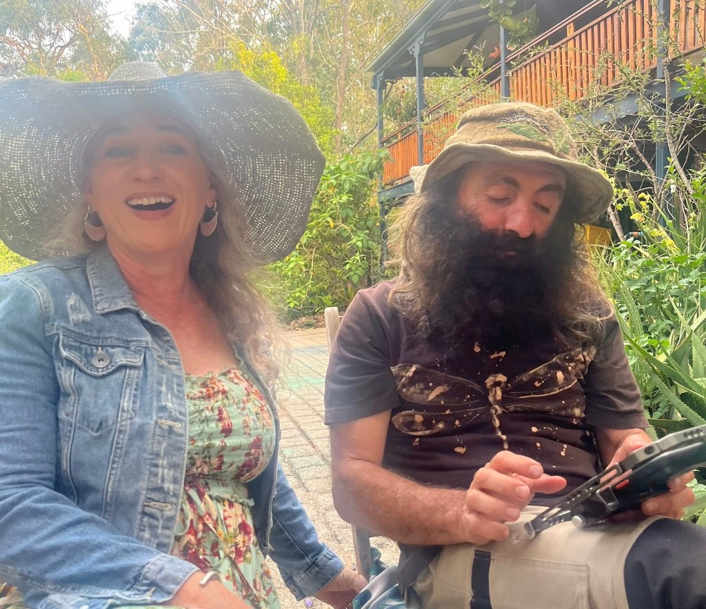 Claire laughing to camera as Costa does something technical with a device on his lap.